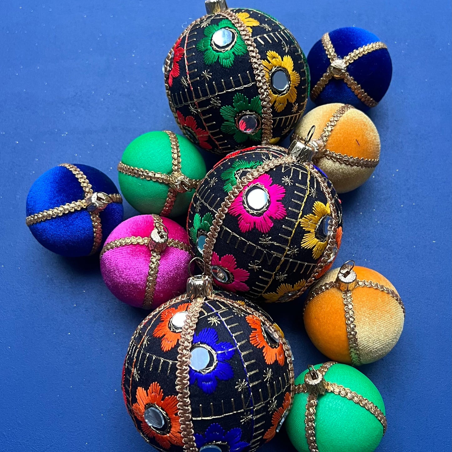 Flatlay of Vimal vibrant black floral handmade baubles, colourful embroidered eco-friendly Christmas ornaments on blue background.