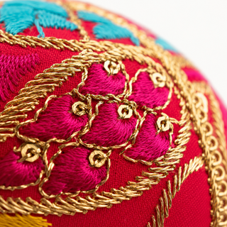 Aditi bauble hanging with soft bokeh lights, showing its hot pink ground and large sweeping gold paisley cordwork with dense magenta satin stitch and multicolour panels.