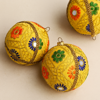 Three yellow decorative balls with floral patterns on a beige background. Aradhya Bauble – sustainable fabric decoration with gold embroidery and colourful floral details on a yellow base, handmade in Britain.