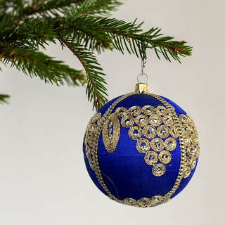 The Arjun Bauble suspended from a fir tree branch, photographed against a clean white background. The deep royal blue bauble features detailed golden embroidery and mirror embellishments, showcasing its intricate craftsmanship.