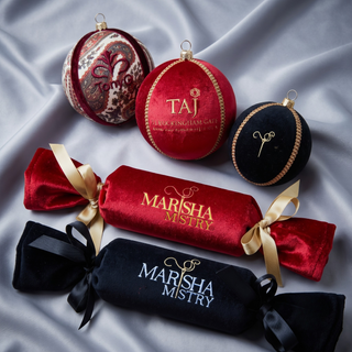 Five luxury gift items arranged on draped silver satin fabric: two round ornaments (one red with gold monogram, one black with gold monogram, and one marbled red/gold ornament), a red velvet cylindrical box tied with a gold ribbon and printed 'MARISHA MISTRY', a black velvet cylindrical box tied with a black ribbon and printed 'MARISHA MISTRY', and a red velvet ornament printed 'TAJ FERRING KING LADY'.