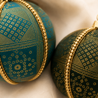 Two decorative Christmas ornaments with intricate gold geometric patterns on teal and dark green fabrics, accented with gold beaded bands, resting on a soft beige fabric background.
