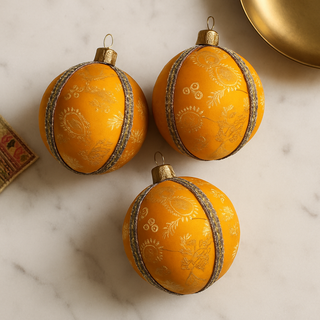 Three yellow ornaments with gold decorative bands on a marble surface. Handcrafted Janvi Bauble in golden brocade fabric with tonal embroidery and antique gold trim, UK-made.