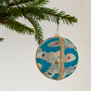 A round Christmas ornament hanging from a green pine branch against a plain light background. The ornament has a turquoise blue and silver patterned body with gold trim and small red and blue accents.