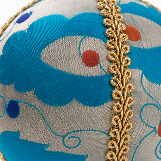 Close-up of a decorative spherical textile object featuring a blue and silver-gray patterned fabric with a vertical gold embroidered vine border and small red and blue dots.