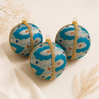 Three round decorative hanging ornaments with a turquoise and white floral pattern, accented by gold trim and gold hanging loops, arranged on a light cream fabric surface with dried grass accents.