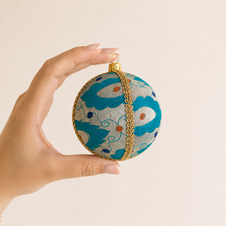A hand holds a round decorative ornament with a turquoise and white abstract pattern, gold trim, and a gold hanging loop against a plain light beige background.