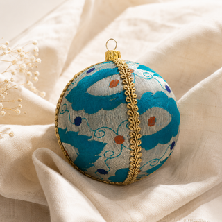A round embroidered Christmas ornament with a turquoise and white floral pattern, a gold decorative trim down the center, and a gold hanging loop, styled on draped cream fabric.
