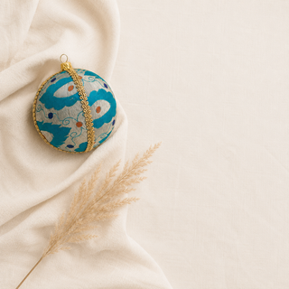 A round decorative ornament with a turquoise and white abstract pattern, accented by a gold chain and gold hanging loop, styled on draped cream fabric with a dried pampas grass stem.