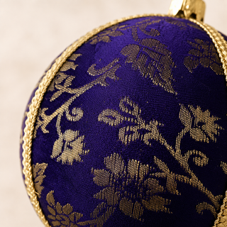 Kaasni bauble hanging with soft bokeh lights, showing its deep purple ground and sweeping gold floral scrollwork brocade with a fine cord seam trim.