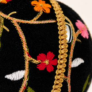 Close-up of a black embroidered textile with floral motifs in red, orange, pink, white, and green, accented by a prominent gold braided cord running vertically through the frame.