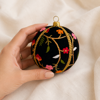 A hand holds a round black embroidered Christmas ornament with gold vine patterns and colorful floral details, set against a soft beige fabric background.
