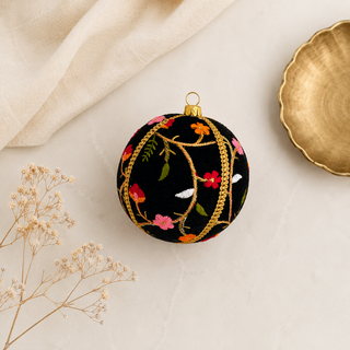 A round black Christmas ornament with colorful floral embroidery and gold vine patterns, styled on a light beige surface with a gold dish and dried flowers.
