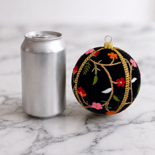 A black embroidered Christmas ornament with gold vine patterns and colorful floral details, shown next to a silver aluminum can on a white marble surface.