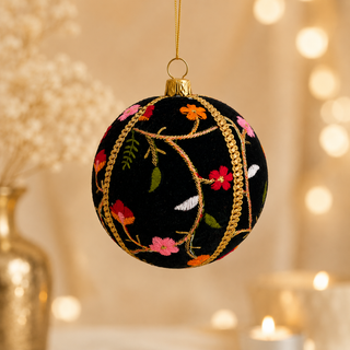 A round black Christmas ornament with gold rope trim and colorful floral embroidery, hanging against a warm bokeh background with blurred lights and a vase.
