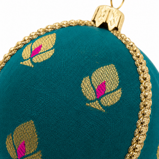 Close-up of a teal green ornament with woven gold floral motifs and bright pink centers, bordered by a gold braided trim and topped with a gold hanging loop.