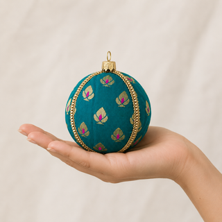 A teal green embroidered Christmas ornament with gold and pink floral motifs and a gold braided trim, held in a person's hand against a light beige marble background.