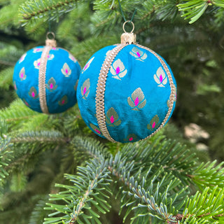 A blue fabric Christmas ornament with a gold metallic trim and pink and green floral pattern, hanging on a green pine tree branch.