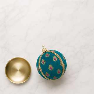 A decorative teal fabric ornament with a gold and pink floral pattern, shown next to its gold-toned metal cap on a white marble surface.