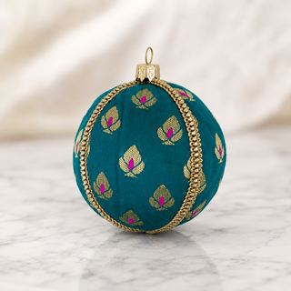 A round teal green decorative ornament with gold woven leaf motifs and pink centers, accented by a gold braided trim and a gold hanging loop, shown on a light marble surface with a soft cream fabric background.