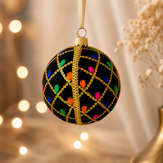 A black Christmas ornament ball with gold chain lattice pattern and colorful embroidered dots, hanging against a warm bokeh background with dried flowers.