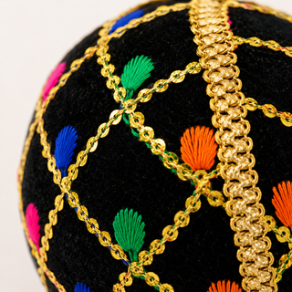 Close-up of a black embroidered decorative ball featuring a gold geometric lattice pattern, colorful faux-fur style accents in green, blue, orange, pink, and white, and a prominent gold braided trim running vertically.
