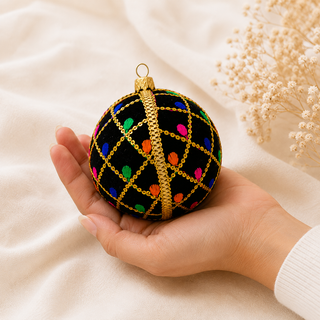 A hand holds a black Christmas ornament decorated with a gold chain lattice pattern and colorful embroidered details, set against a soft beige fabric background with dried flowers.