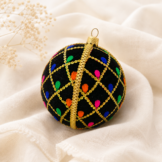 A round black embroidered Christmas ornament with gold chain-like lines, colorful embroidered dots, and a gold hanging loop, styled on draped cream fabric with small dried flowers in the corner.