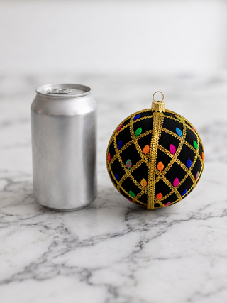 A black Christmas ornament with a gold lattice pattern and colorful hanging lights, shown next to a silver aluminum can on a white marble surface.