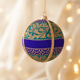 A spherical decorative ornament hanging from a gold string, featuring a teal and purple patterned body with a wide purple horizontal band and gold rope details, set against a softly blurred warm light background.