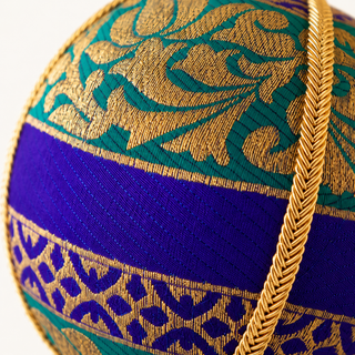 Close-up of a decorative spherical object featuring a rich color scheme of royal blue, emerald green, and metallic gold. The surface is detailed with intricate woven patterns and a braided gold cord running vertically through the frame.
