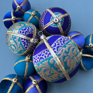 A cluster of decorative spherical ornaments in shades of royal blue, turquoise, and teal, decorated with metallic gold patterns and wrapped with gold rope details, set against a solid blue-gray background.