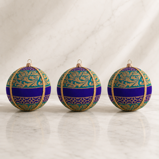 Three decorative spherical ornaments with a rich purple and teal floral pattern, accented by gold trim and hanging loops, displayed on a light marble surface.