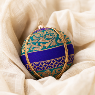 A decorative spherical ornament with a purple and teal patterned fabric body, accented by gold metallic embroidery and rope trim, resting on draped cream fabric.