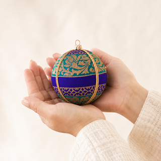 A person's hands holding a decorative spherical ornament with a teal and purple floral pattern, a thick royal blue horizontal band, and gold metallic accents, set against a soft beige background.