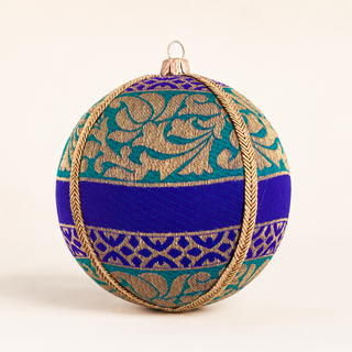 A spherical Christmas ornament decorated with royal blue fabric, teal and gold patterned brocade, and a braided gold rope border, shown on a plain light background.