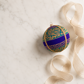 A decorative spherical ornament with an intricate gold and teal floral pattern, a bold purple horizontal band, and a gold hanging loop, styled on a white marble surface with a beige ribbon.
