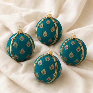 Four decorative spherical ornaments with a teal fabric pattern, gold trim, and gold hanging loops, arranged on draped cream fabric.