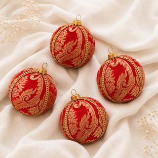 Four red Christmas ornaments with gold damask patterns and gold trim, arranged on a draped cream fabric background with small dried flowers.