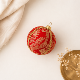 A red Christmas ornament with an ornate gold damask pattern and gold trim, styled on a light beige background with a cream fabric corner and dried flowers.