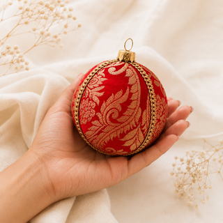 A hand holds a round red Christmas ornament decorated with an ornate gold damask pattern and a gold hanging loop, set against a soft beige fabric background with dried baby's breath flowers.