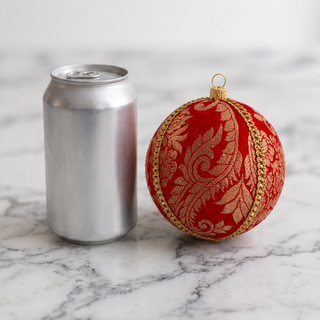 A red Christmas ornament with gold ornamental patterns and a gold hanging loop, shown next to a silver aluminum can on a white marble surface.