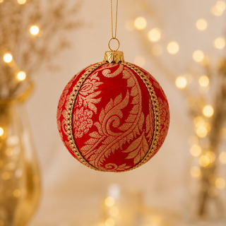 A red velvet Christmas ornament with intricate gold damask embroidery and gold trim, hanging against a warm, softly blurred background with bokeh lights.