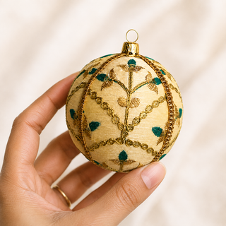 Lalit fabric bauble styled on linen, a 10cm handmade ornament with delicate gold floral embroidery with teal thread accents and a metallic gold trim.