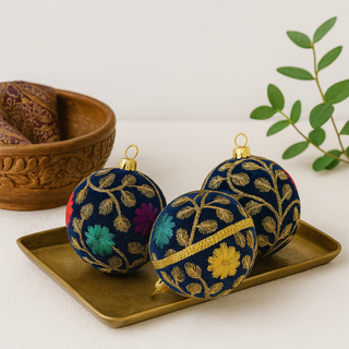 Decorative Mahir bauble with floral embroidery on a gold tray with a wooden bowl and green leaves in the background. Mahir Bauble – sustainable fabric decoration with jewel-toned embroidered details on navy velvet, handmade in Britain