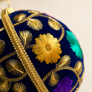 Mahir bauble hanging with soft bokeh lights, showing its deep navy ground and fine gold fern-leaf embroidery with scattered pink, purple and teal florals.