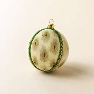 Kamla bauble hanging with soft bokeh lights, showing its soft cream (also in black) ground and a delicate gold floral motif with green accents and a green-and-gold cord trim.