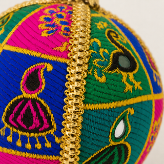 Close-up of a round embroidered ornament or decorative ball with a gold metallic chain down the center. The fabric features multiple bright colors including blue, green, pink, and yellow in geometric and peacock-inspired patterns.