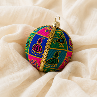A round decorative ornament with a colorful patchwork pattern of pink, blue, and green sections, featuring embroidered peacock motifs and a gold braided center band, resting on draped cream fabric.