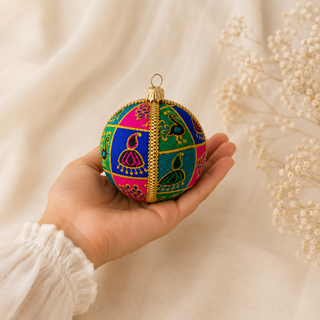 A hand holds a colorful embroidered Christmas ornament ball with a gold top, set against a soft cream fabric background with dried baby's breath flowers.